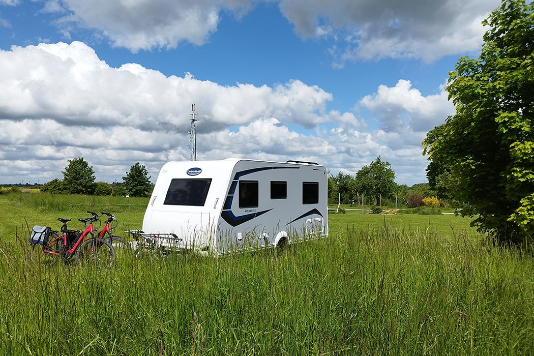 Witte caravan met twee fietsen ervoor op een grasveld met hoog gras op camping MiO in Ottendorf bij Chemnitz. Op de achtergrond bomen en een zendmast.