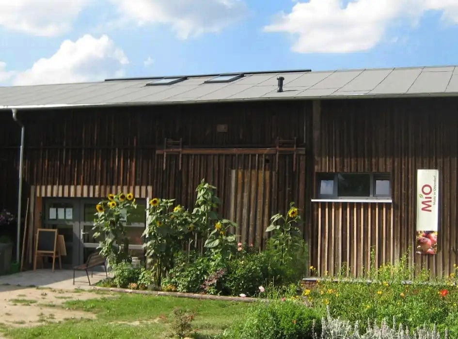 Holzfassade mit Blumenbeet und Sonnenblumen vor dem Eingangsbereich des Campingplatzes MiO in Ottendorf bei Chemnitz. An der Wand ein Schild mit der Aufschrift 'MiO'.