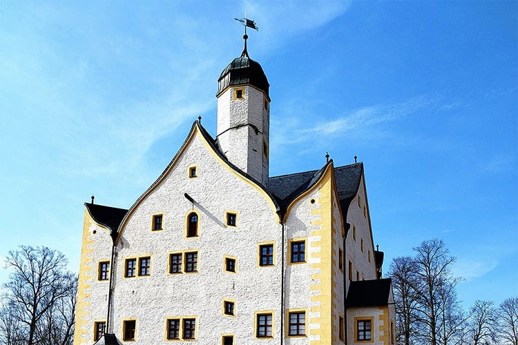Frontale Ansicht des Wasserschlosses Klaffenbach mit markantem Turm, weißen Fassaden und gelben Fensterrahmen vor blauem Himmel.