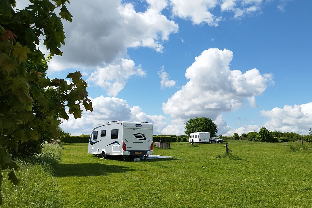 Twee caravans op een uitgestrekt grasveld op camping MiO in Ottendorf bij Chemnitz. Op de voorgrond een boom met groene bladeren, erboven een bewolkte lucht.