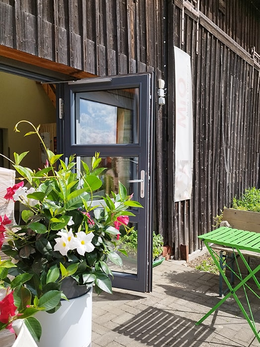 Open glazen deur aan een houten gebouw op camping MiO in Ottendorf bij Chemnitz. Ernaast een bloembak met rode en witte bloemen en een groene klaptafel met bank.