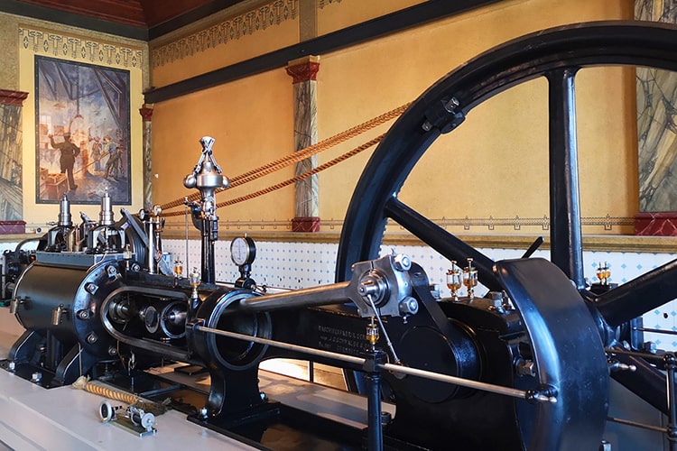 Detailansicht einer historischen Dampfmaschine in einem Museumsraum mit dekorativer Wandgestaltung und Gemälde im Hintergrund.
