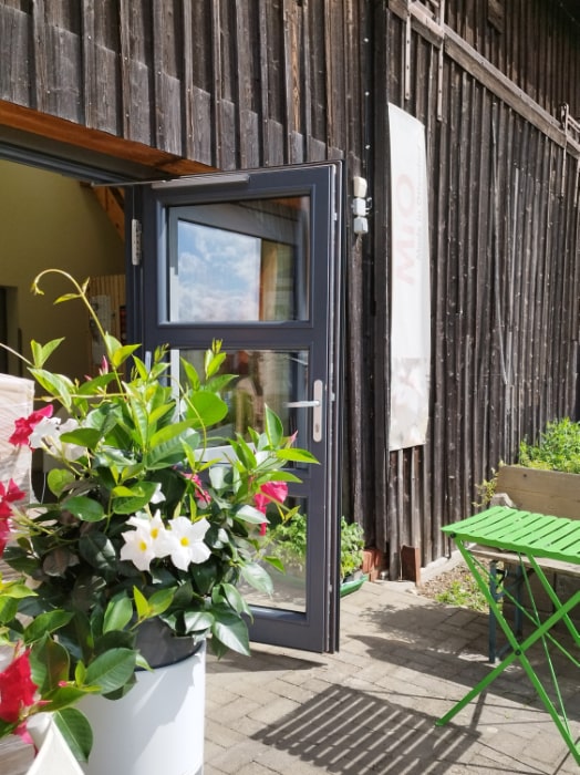 Offene Glastür an einem Holzgebäude auf dem Campingplatz MiO in Ottendorf bei Chemnitz. Daneben ein Blumenkübel mit roten und weißen Blüten sowie ein grüner Klapptisch mit Bank.