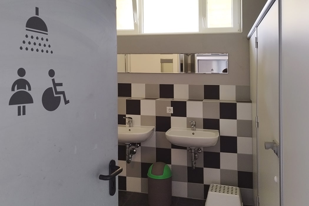 Ingang van een sanitaire ruimte met twee wastafels, spiegel, afvalbak en opstapje op camping MiO in Ottendorf bij Chemnitz. Op de open deur staan pictogrammen voor douche, vrouwen en rolstoelgebruikers.