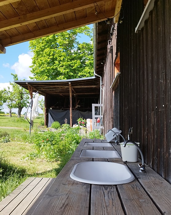 Meerdere spoelbakken met kranen aan een houten werkblad buiten een gebouw op camping MiO in Ottendorf bij Chemnitz. Op de achtergrond een overdekte zitplek en bomen.