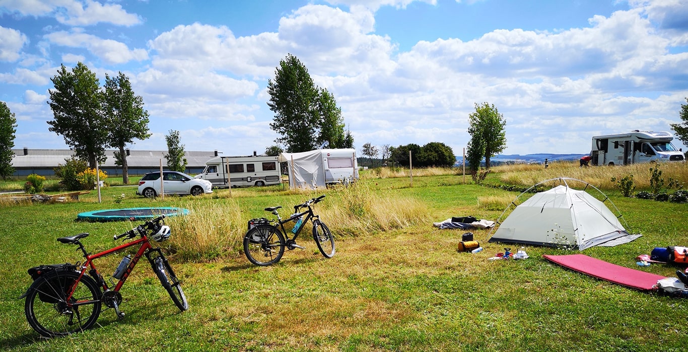 Tent, fietsen, kampeeruitrusting en voertuigen op een open grasveld op camping MiO in Ottendorf bij Chemnitz. Op de achtergrond bomen, campers en een weids uitzicht over het landschap.