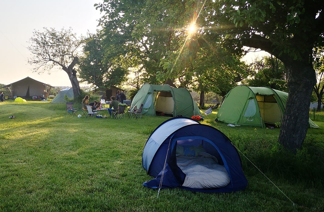 Tenten en kampeerstoelen onder bomen op een grasveld op camping MiO in Ottendorf bij Chemnitz. Op de voorgrond een open kleine tent, op de achtergrond meer tenten en een vast tenthuis.